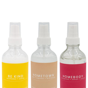 Four frosted glass aroma spray bottles with white lids. The bottles are labelled with different colored labels indicating a different scent. There is a neutral background with a beige vase in the background, the vase has some greenery in it.