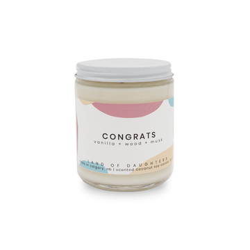 A glass jar candle with a white lid and a white, pink and blue label that reads Congrats, vanilla + wood + musk on a white background.