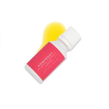 A small white glass bottle with a dark pink label that reads Homebody, bergamot + lemon + Vanilla rests upon a small pool of fragrance oil