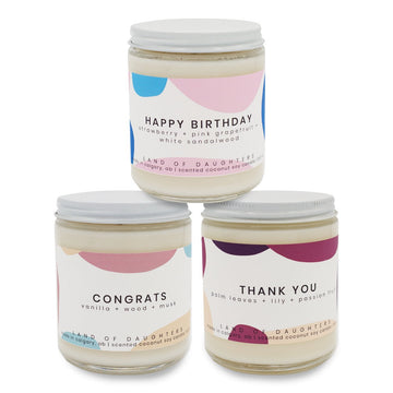 Three soy candles in glass jars with white lids. The candles have white labels across them with different colored abstract shapes. They are sitting in a beige basket on a white background.