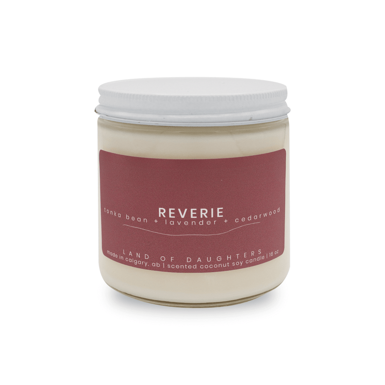 16oz Glass Jar candle with white lid and a dark purple label that reads Reverie, tonka bean + lavender + cedarwood is photographed on a white background.