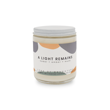 8oz Glass Jar candle with white lid and a light orange and blue label that reads A Light Remains, cedar + amber + musk is photographed on a white background.