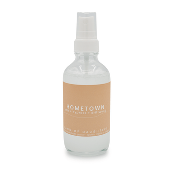 A glass spray bottle filled with liquid and a beige label that reads Hometown, salt + cypress + driftwood on a white background