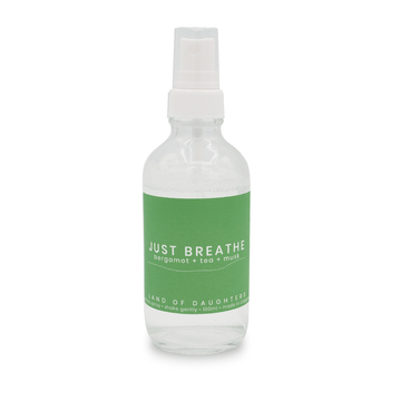 A glass spray bottle filled with liquid with a green label that reads Just Breathe, bergamot + tea + musk photographed on a white background