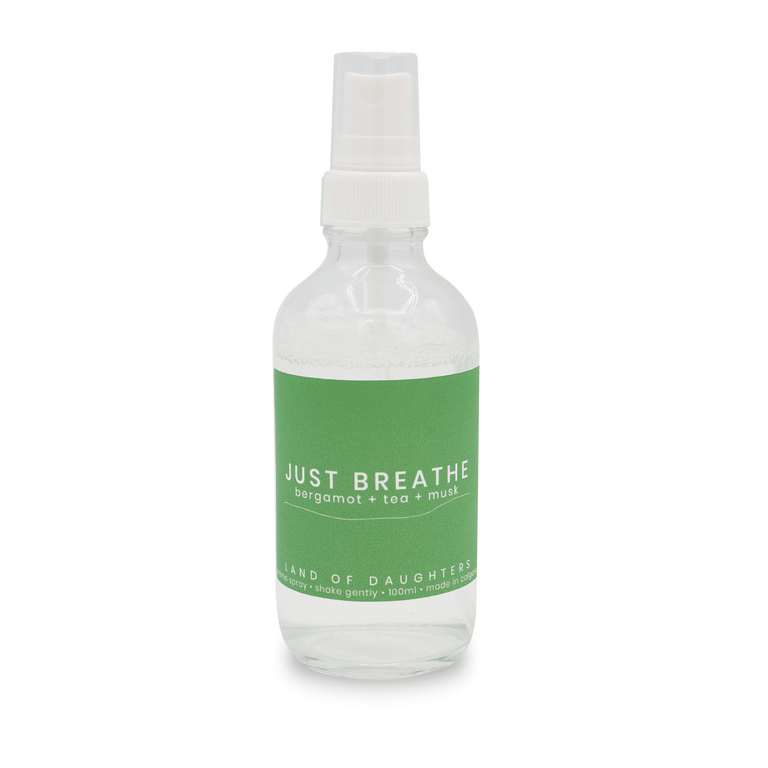 A glass spray bottle filled with liquid with a green label that reads Just Breathe, bergamot + tea + musk photographed on a white background