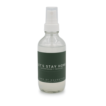 A glass spray bottle filled with liquid with a dark green label that reads Let's Stay Home, sandalwood + lavender is photographed on a white background.