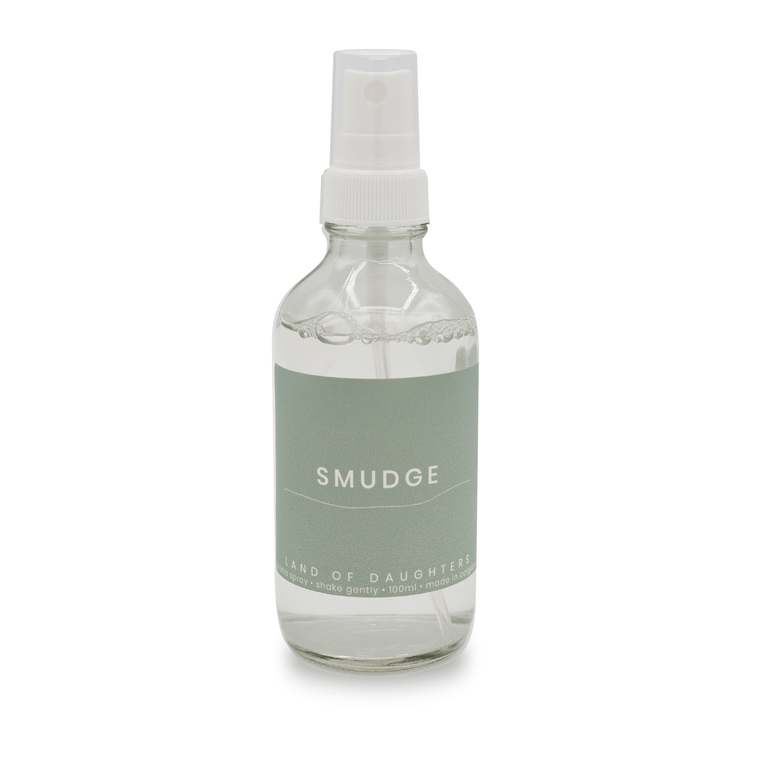 A glass spray bottle filled with liquid with a light green label that reads Smudge is photographed on a white background.