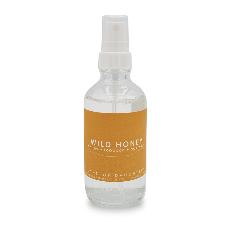 A glass spray bottle filled with liquid with a light orange label that reads Wild Honey, honey + tobacco + oak moss is photographed on a white background.