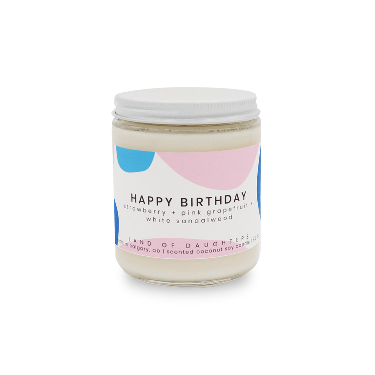 a glass jar candle with a pink and blue label that reads Happy Birthday, strawberry + pink grapefruit + white sandalwood on a white background