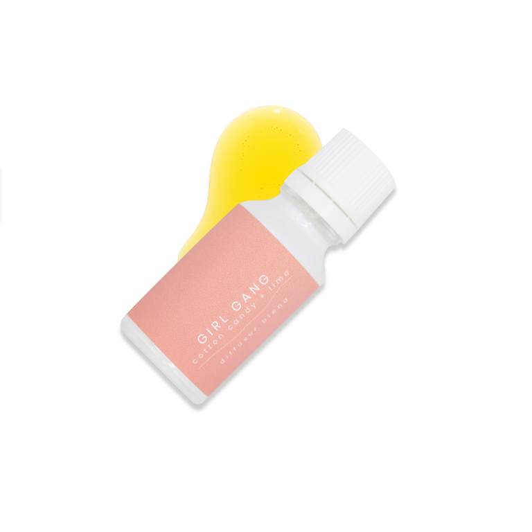 Small white glass bottle with white lid and a light pink label that reads Girl Gang, cotton candy + lime resting on a small pool of fragrance oil