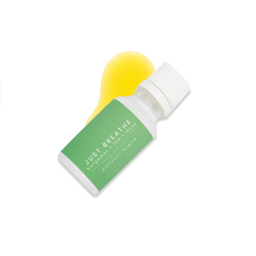 A small white glass bottle with white lid and a green label that reads Just Breathe, bergamot + tea + musk rests upon a small pool of fragrance oil on a white background.