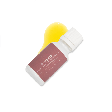 A small white glass bottle with white lid and a dark purple label that reads Reverie, tonka bean + lavender + cedarwood rests upon a small pool of fragrance oil on a white background.