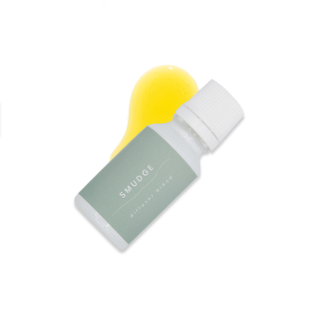 A small white glass bottle with white lid and a light green label that reads Smudge rests upon a small pool of fragrance oil on a white background.