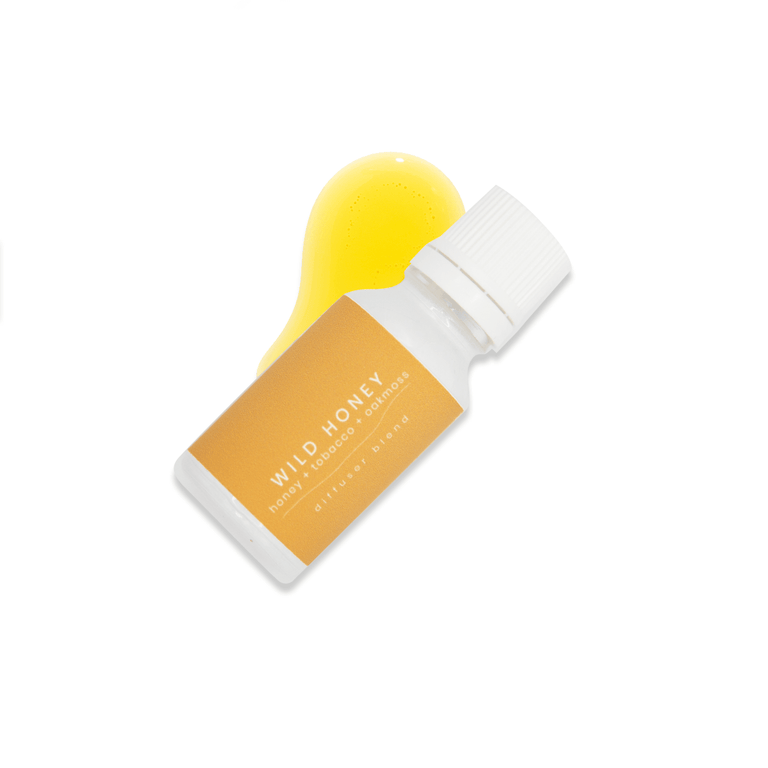 A small white glass bottle with white lid and a light orange label that reads Wild Honey, honey + tobacco + oak moss rests upon a small pool of fragrance oil on a white background.