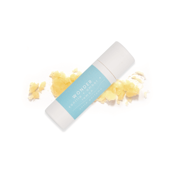 A white paperboard tube with a light blue label that reads Wonder, vanilla + amber + lemon resting upon a smear of wax below on a white background.