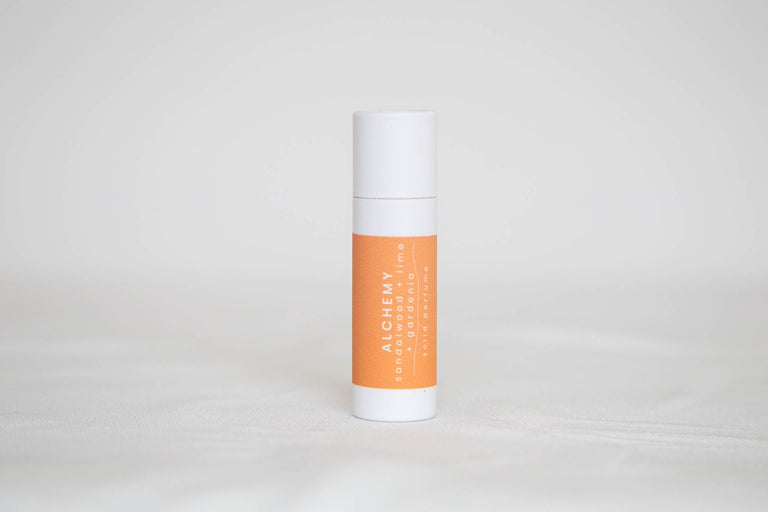 A white paperboard tube of solid perfume with a light orange label that says alchemy, sandalwood, lime and gardenia. On a white background.